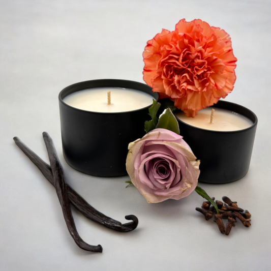 Black jars of Ophelia hand poured soy candles with rose, carnation, vanilla, and sandalwood next to flowers and spices on a white background.